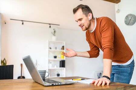 Video chat online, video call, zoom. A young bearded guy speaks using a laptop. He is standing, looking at the camera, explaining something and smiling.の写真素材