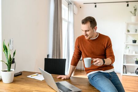Remote communication using video calls. A young handsome guy is talking using a laptop, he is sitting at the edge of the table at ease and with a cup of coffee in his hand and smiling into the webcamの写真素材