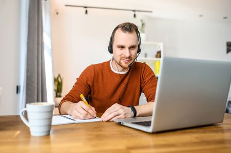 Work from home. A guy is using headset and laptop for work, he notes information in notebookの写真素材