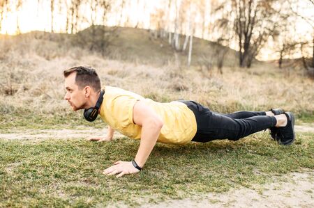 Young athletic guy with headphones has workout outdoors. He is doing push ups on the ground. The concept of self-discipline and a healthy lifestyle.の写真素材