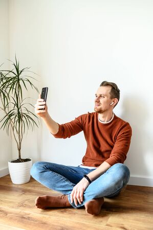 The guy takes a selfie, speaks via video. A young guy sits at home on the floor in the living room, takes pictures of himself on the front camera of the phone and smilesの写真素材