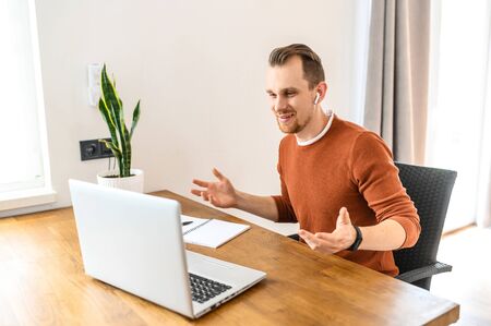 Video call. A young positive guy in casual clothes is using a laptop and airpods for online communication. He sits at the table, looks at the laptop screen and talks. Back viewの写真素材