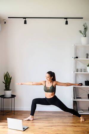Yoga video tutorials. Girl training at home, practicing yoga and watching videos on laptop in living room. She is doing warrior asana. Sport at home conceptの写真素材