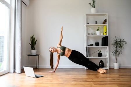 Get in shape. Young positive woman learn yoga from video tutorials. She looks at the laptop screen and repeats exercises and asanas.の写真素材