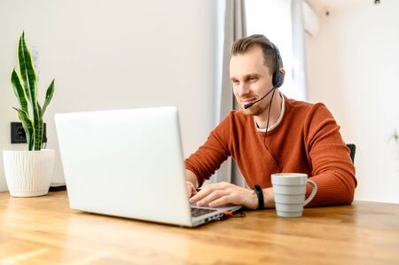 The guy uses hands-free headsets to work from home. He sits at a table with a laptop and speaks online. Remote work conceptの写真素材