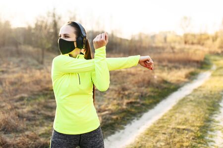 Get in shape during quarantine. Athletic woman is doing cardio workout outdoors. She in a medical mask doing stretching on the grass. Spending time with healthy benefit during coronavirus epidemiaの写真素材
