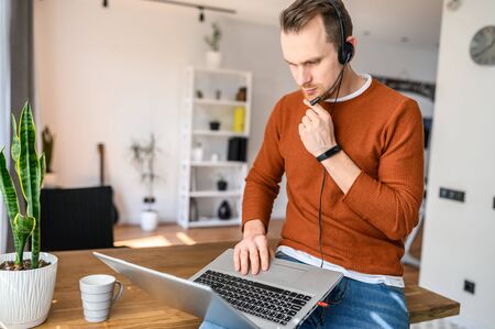 A freelancer guy works from home. Guy with a hands-free headset in the sits on the edge of table, looks at the laptop screenの写真素材
