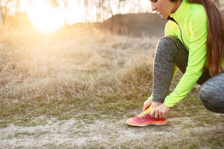Preparations for the morning jogging. Young woman in headphones and in bright sportswear ties up her shoelaces and prepares to run. Close-up pictureの写真素材