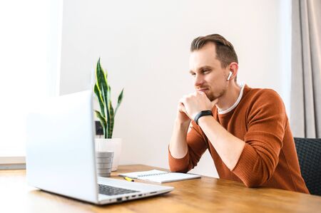A portrait of concentrated guy using laptop and headphones for work. He looks at screen thoughtfullyの写真素材