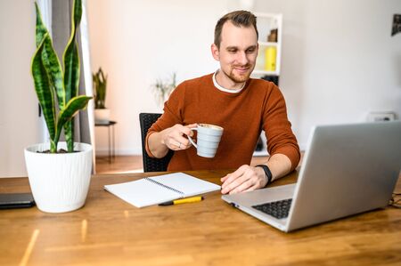 Morning coffee on the workplace. Handsome positive guy using laptop for work, he sits with cup of coffee in hands and looks in screen with a smileの写真素材