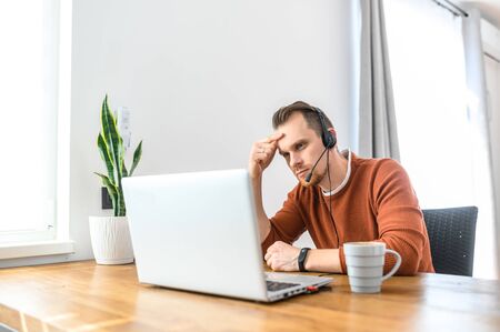 A guy call center operator, support service operator at home office. He uses a headset to talk sits...
