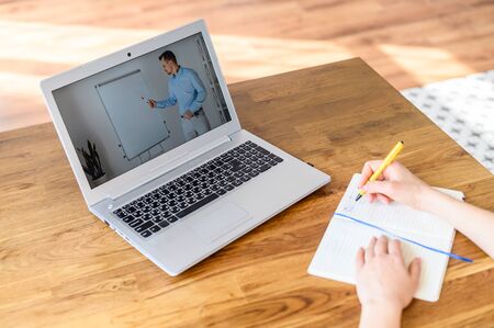 Female hands writing a syllabus in notebook, a laptop with a online lecture on the table. Concept of...