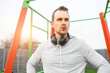 An athletic guy in hoodie and with headphones on the outdoor sports ground with a horizontal bars