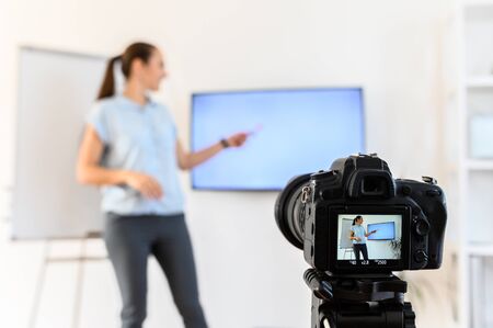 A woman is recording video classes at home for distance study, she uses flip chart and monitor. Camera on tripod on foreground. Woman is out of focusの写真素材