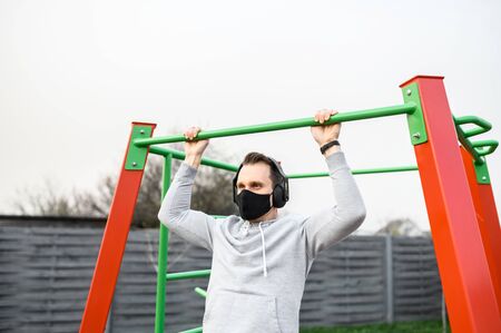 A young man is working on a horizontal bar outdoors during quarantine, he is doing pull-ups in...