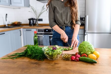 Hands of woman are cutting cucumber on wooden cutting board, face is not visible. Fresh vegetables and greens on the table near for cooking salad. Healthy food conceptの写真素材