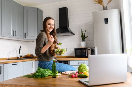 A young woman eats a fresh salad in the kitchen and looks with interest at the laptop screen. She is watching series, movies. Vegetables on the tableの写真素材