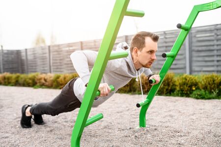 A guy in a hoodie is doing angle push-ups outdoors. Upper body workout on sports ground