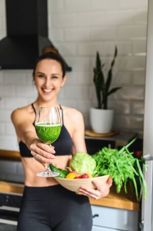 Body detox. Young attractive woman in sportswear in the kitchen with a glass of detox smoothie and a basket of vegetables in her handsの写真素材