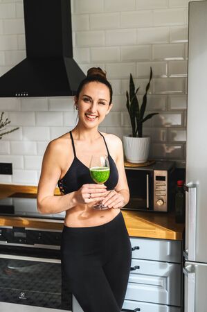 A glass of vegetable smoothie in the hands of a young healthy woman, she is in sportswear standing in the kitchen and smiling. Vertical photoの写真素材