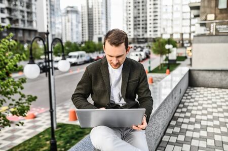 Work outdoors. A young man in smart casual clothes uses a laptop for work, a skyscraper in the...