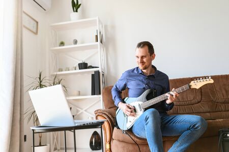 Online studying. A young man is watching video tutorial, video classes how to play guitar, he sits on couch with electric guitar and combo amp nearの写真素材