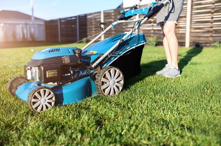 Close-up blue push lawn mower on a green grass, man on a background. A man mows grass