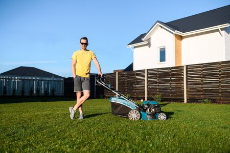 Portrait of guy with a lawn mower on a back yard of private house, he is in contemporary sunglasses and in yellow casual t-shirt looks at camera and smiles. Care about backyardの写真素材