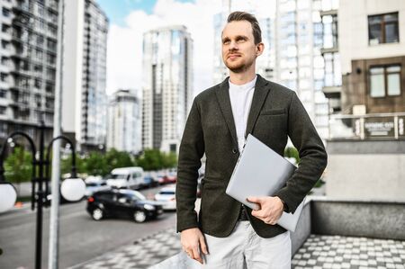 A young businessman among the skyscrapers in the city, he holds a laptop in his hand and looks forward with confidenceの写真素材