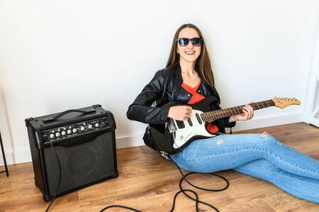 A young beautiful woman is playing an electric guitar using a combo amplifier, she is in a black leather jacket and in sunglasses sits on the floor at homeの写真素材