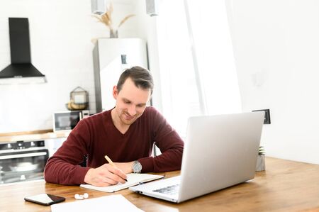 Male student writes a lecture in a notebook at home. He uses a laptop to watch a lecture.の写真素材