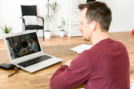 Remote work. A young guy uses laptop for video call, he sits at the kitchen table and looks in webcam, his coworkers on the screen. Back viewの写真素材