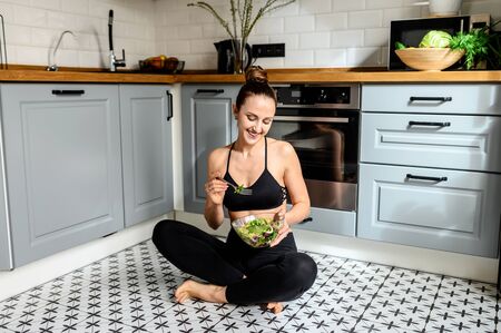 The concept of a healthy nutrition and healthy lifestyle. Young woman with a bowl of salad sitting in the kitchen on the floor, eating salad and smilingの写真素材