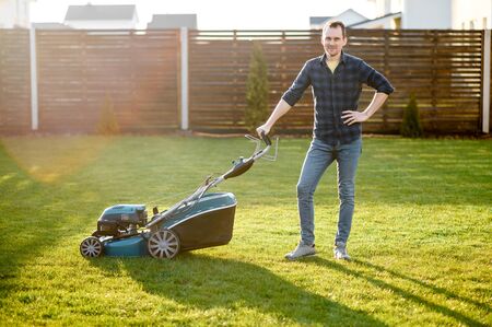 Portrait of guy with a lawn mower on a back yard of private house, he stands and smiles. Care about backyardの写真素材