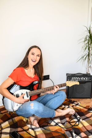 Vertical photo of young posive girl with electric guitar, she sits on the plaid in cozy homely atmosphere, combo amp nearの写真素材