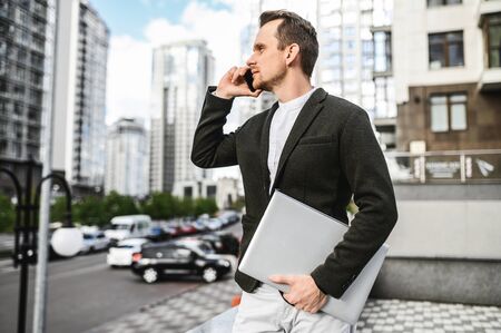 A young businessman in smart casual clothes is talking on the phone amid the cityscape, he is holding a laptop in his handsの写真素材