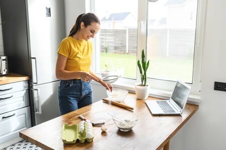 Cook at home. A young woman in casual clothes in the kitchen is going to bake a cake, cookies, she is sifting flour on the tableの写真素材