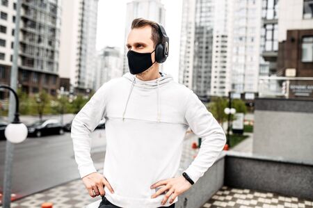 Sports lifestyle. Young athletic man in sportswear in a medical mask with headphones among city skyscrapersの写真素材