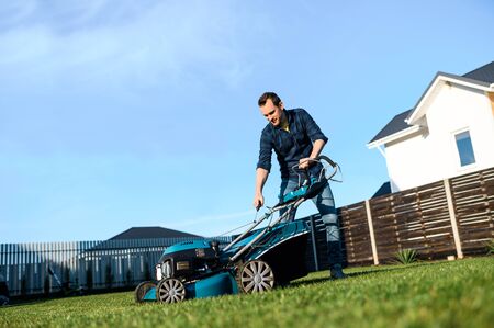 A young man is going to mow the lawn, he starts a push lawn mower. A guy in casual plaid shirt and in jeansの写真素材