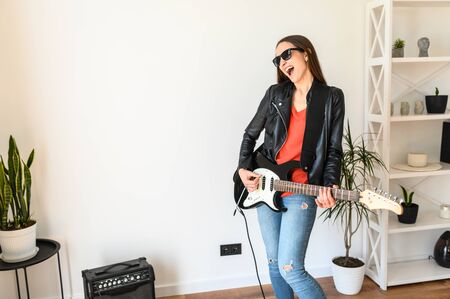 Home leisure with fun. A young beautiful woman in sunglasses and in leather jacket is playing an electric guitar indoors, she is smiling happily. Hobby concept, music talentの写真素材
