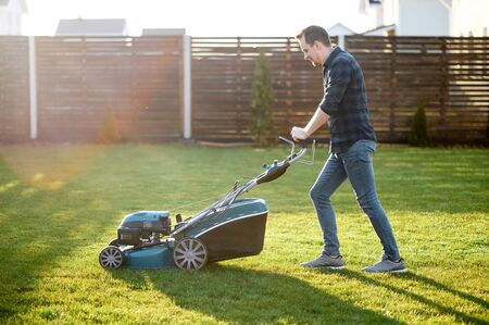 Spring mowing the lawn. Young man in a plaid shirt and jeans mows grass with a lawn mower in the backyard. Side viewの写真素材