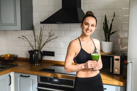 A glass of vegetable smoothie in the hands of a young healthy woman, she is in sportswear standing in the kitchen and smilingの写真素材