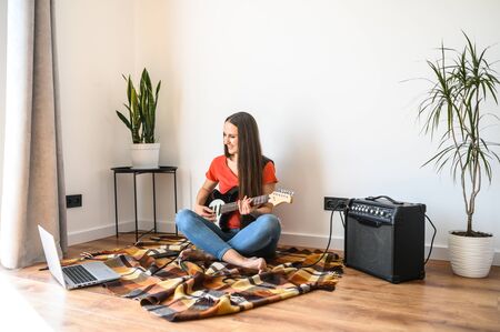 Hobbies and leisure activities during quarantine. Online training, classes. A young woman watches a video lesson on playing the electric guitar, she sits on a cozy plaid with a guitar, combo amp nearの写真素材