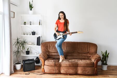 Fun at home on self-isolation. A young woman with an electric guitar in her hands dances and sings on a sofa, she using a sombo amplifierの写真素材
