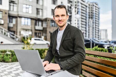 A young happy man in smart casual uses laptop for remote work outdoors. He sits on the bench among cityscape, looking at camera with smileの写真素材