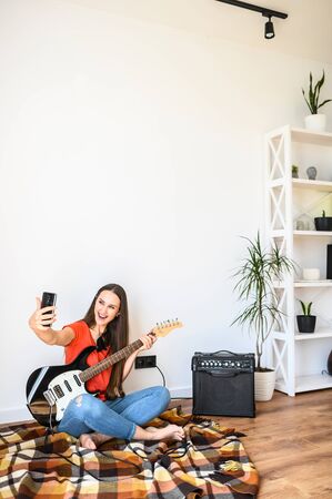 Young positive woman makes selfie on the phone with an electric guitar at home, combo amp near. She is sitting on the floor on a cozy plaid, is looking at the smartphone camera and smiling.の写真素材