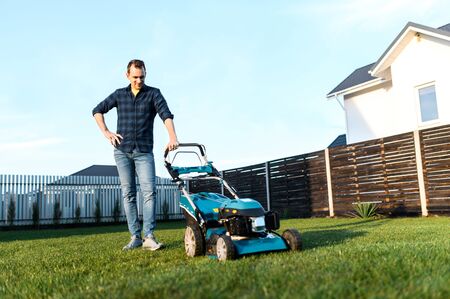 Portrait of guy with a lawn mower on a back yard of private house, he stands and smiles. Care about backyardの写真素材