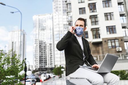 A young businessman uses a laptop to work outdoors during the coronavirus epidemic. He is sitting on a bench among the skyscrapers with a medical mask on his face and talking on the phoneの写真素材