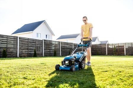 A handsome guy is mowing a lawn with a gasoline lawn mower and smiles, he is in sunglasses. Care about backyardの写真素材
