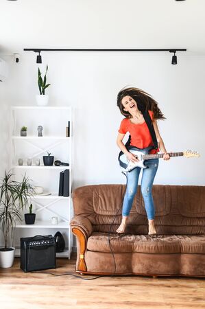 A girl with an electric guitar has a fun at home, she jumps on couch and laughsの写真素材